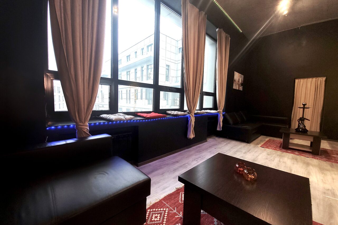 Арендовать Элегантный лофт с хорошим звуком и балконом. Лофт-пространство с черной кожаной мебелью, низким черным столом, большими окнами с бежевыми шторами, светлый пол, стены темного цвета, в углу — кальян, на стене картина, есть декоративная подсветка.