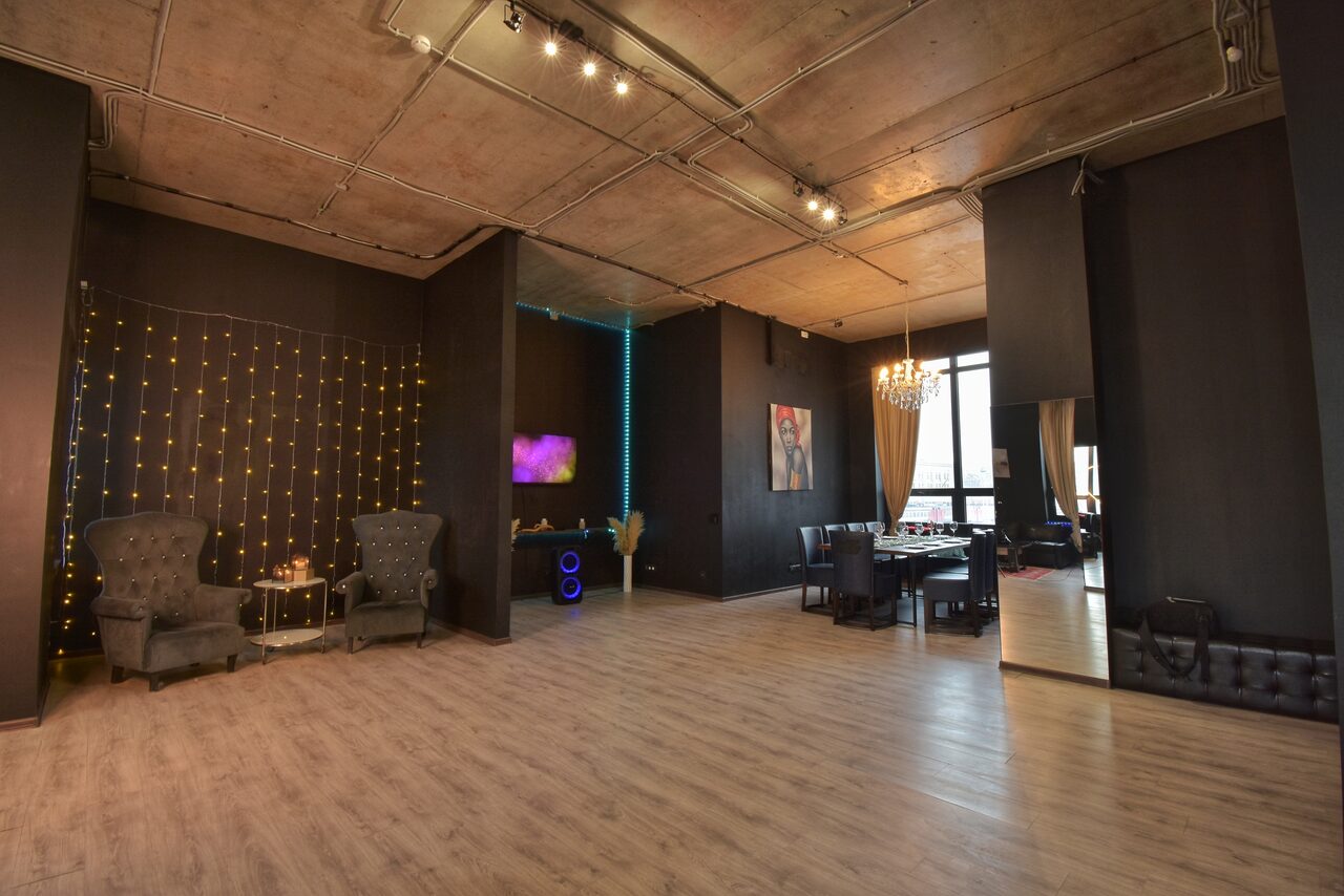 Арендовать Элегантный лофт с хорошим звуком и балконом. Просторное современное помещение с бетонным потолком и светлым деревянным полом. Чёрные стены с гирляндами, зона отдыха с двумя креслами, столик, обеденный стол со стульями. Большое окно с занавесью, зеркало, подсветка. Декорации на стенах.