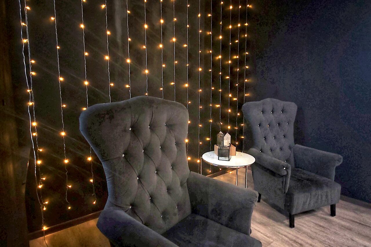 Арендовать Элегантный лофт с хорошим звуком и балконом. Два темных мягких кресла в комнате с деревянным полом. Между креслами стоит круглый белый стол с декором на поверхности. Темная стена задрапирована нитями теплых огоньков.