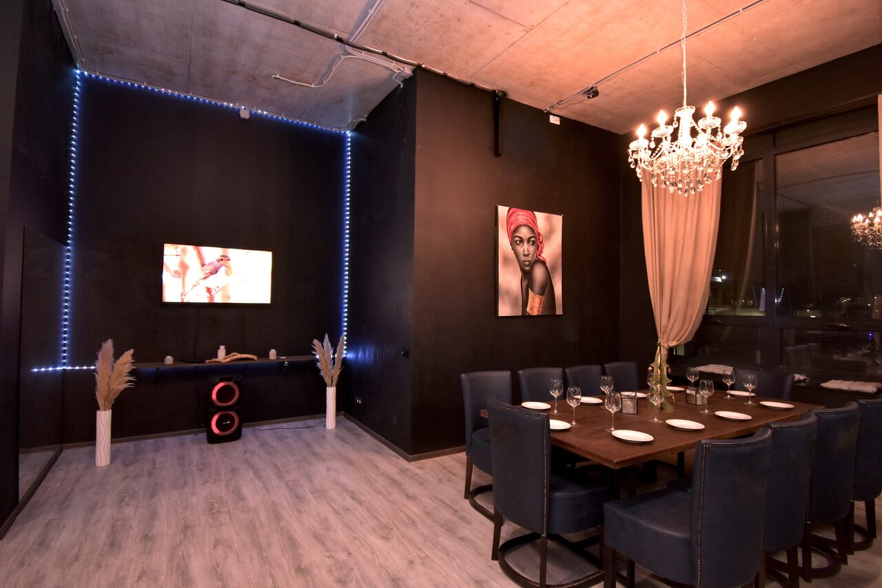 Арендовать Элегантный лофт с хорошим звуком и балконом. Темная комната с высоким потолком и бетонными элементами. В центре большой деревянный стол с синими стульями. На стене картина и телевизор. В углу динамики. Хрустальная люстра и светодиодное освещение. Серые полы, большие окна с занавесками.