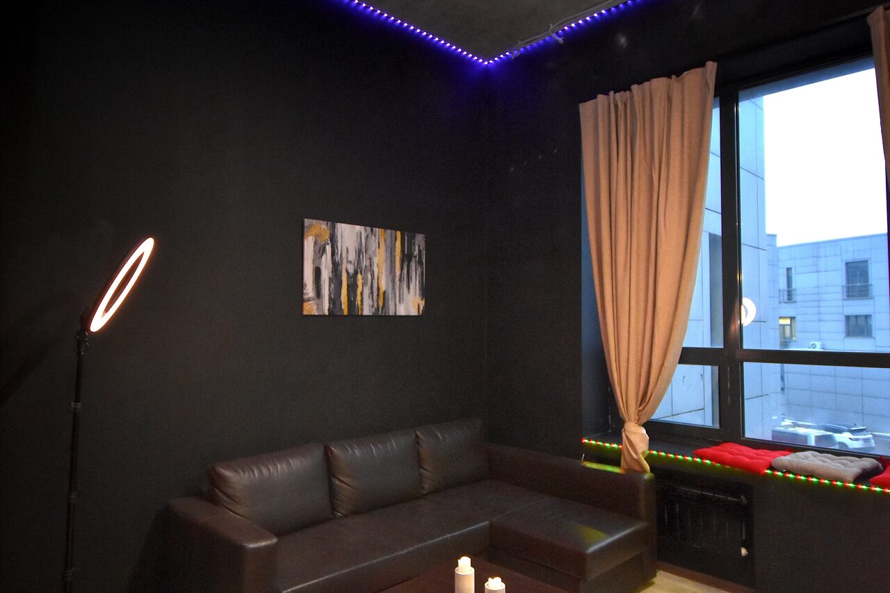 Арендовать Элегантный лофт с хорошим звуком и балконом. Гостиная с тёмными стенами и угловым диваном из чёрной кожи. Полотно абстрактного искусства на стене. Окно с бежевыми шторами. Светодиодное и кольцевое освещение. Стеклянный журнальный столик. Вид на здание через окно.