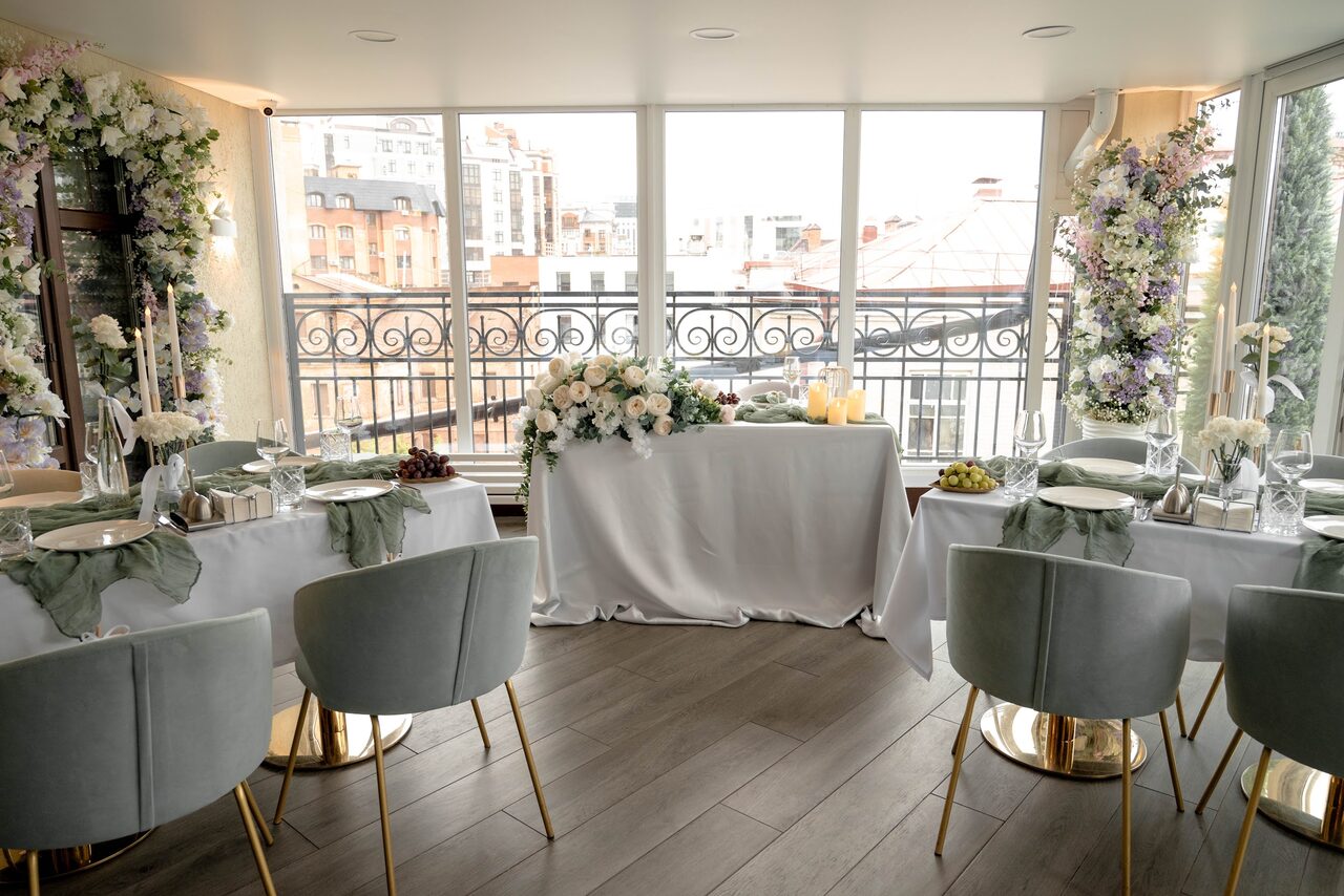 Арендовать Ресторан Небо Казани. Пространство с панорамными окнами, видом на город, три накрытых стола с белыми скатертями, декоративные свечи и цветочные композиции, серые мягкие стулья с золотыми ножками, пол из темного дерева, украшенные цветами стены.