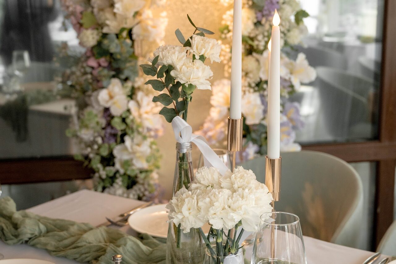 Арендовать Ресторан Небо Казани. Элегантный стол сервирован белыми свечами в подсвечниках, букетом белых цветов в стеклянной вазе и светлой скатертью. На заднем плане стенка с аркой из цветов. Мягкое освещение.