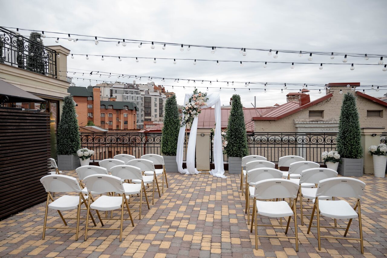 Арендовать Ресторан Небо Казани. Открытая терраса с выложенным плиткой полом, рядами белых стульев с золотыми ножками, аркой украшенной тканью и цветами. По периметру террасы расположены высокие растения в горшках. Над пространством натянуты гирлянды с лампочками.