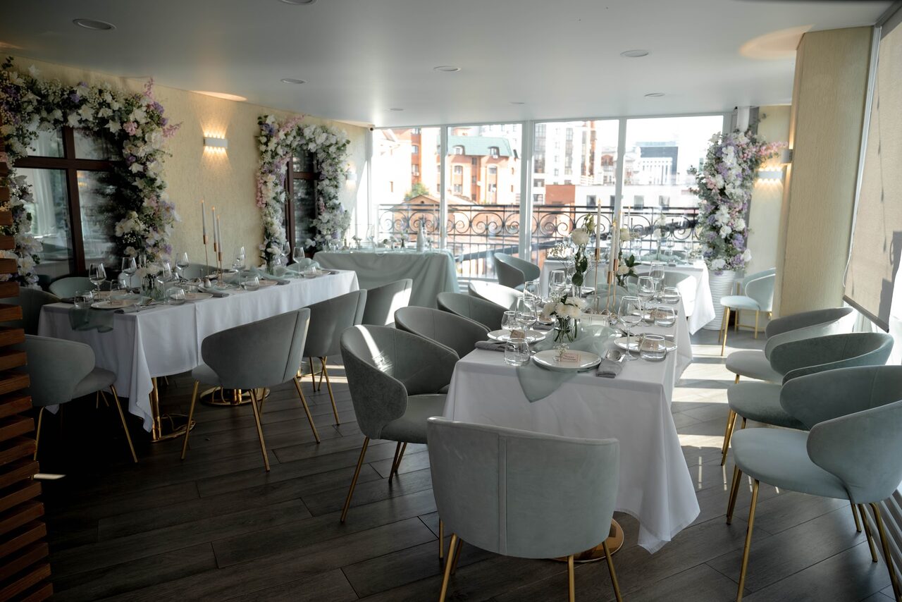 Арендовать Ресторан Небо Казани. Банкетное пространство с большими окнами, накрытые белыми скатертями столы, серые стулья с золотыми ножками, сервировка для гостей. Декор из цветов на стенах, деревянный пол, светлые стены, дневное освещение.