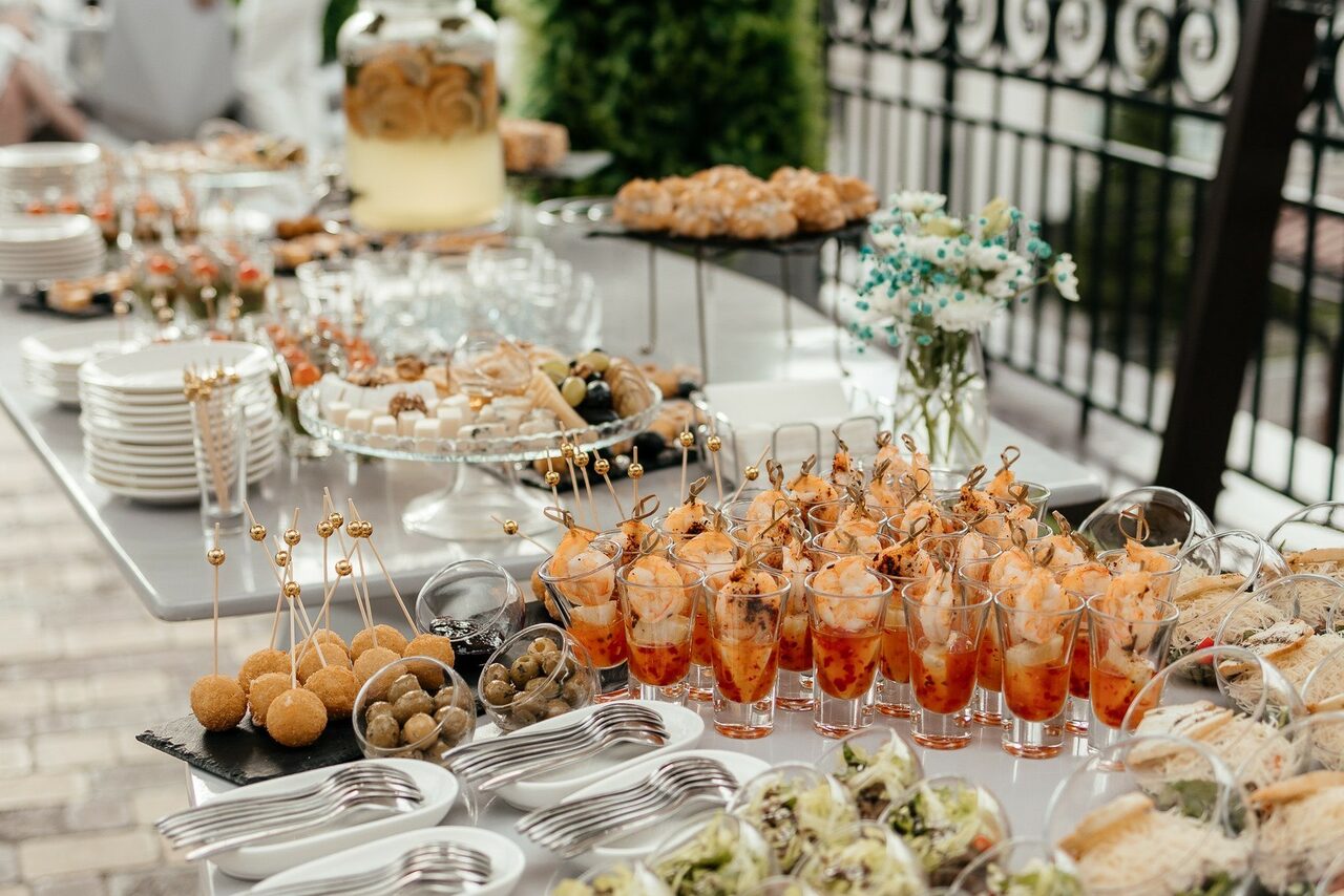Арендовать Ресторан Небо Казани. Настольная сервировка в наружном пространстве: длинный стол с закусками в бокалах и на шпажках, канапе, оливки. Есть стеклянные стаканы, блюда из металла и стекла. На заднем плане видны декоративные растения и кованая ограда.