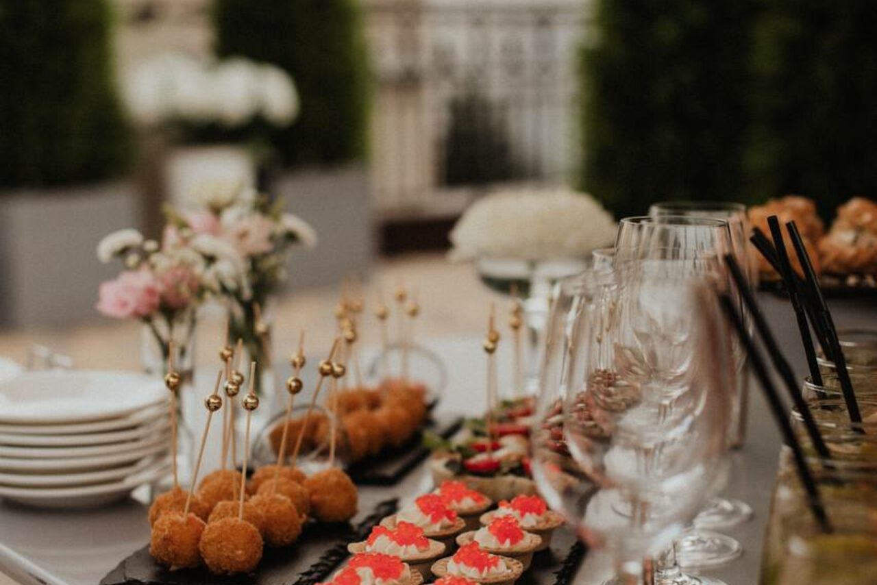 Арендовать Ресторан Небо Казани. Стол с закусками и бокалами на открытом воздухе. На столе тарелки с канапе и закусками, цветочная композиция, пустые бокалы на высоких ножках, коктейльные трубочки. Фон размытый, с элементами зелени и стены.