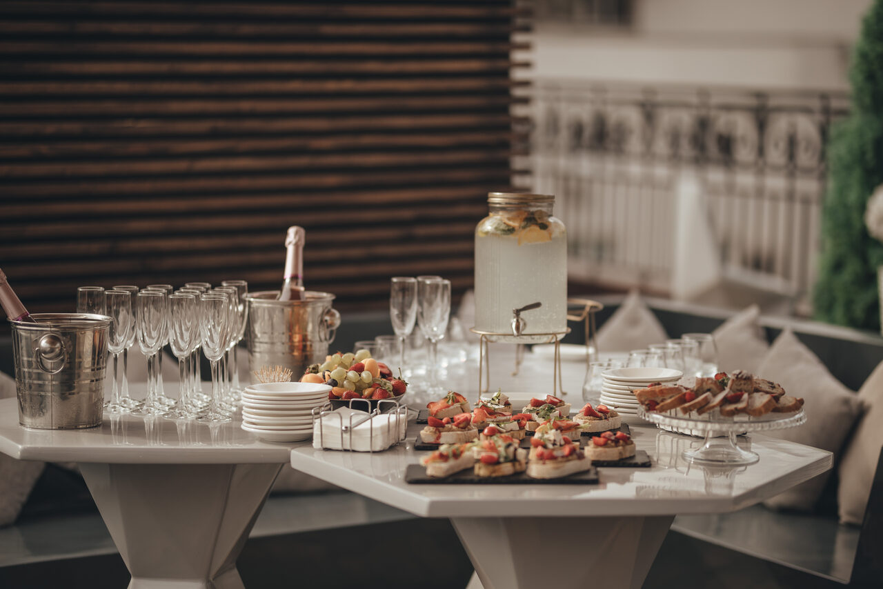 Арендовать Ресторан Небо Казани. Настольное пространство с закусками, напитками в стаканах и кувшине, бокалами для шампанского, металлическим ведром с бутылками. Белые и серые тона, диваны с подушками на заднем плане, деревянная панель на стене.