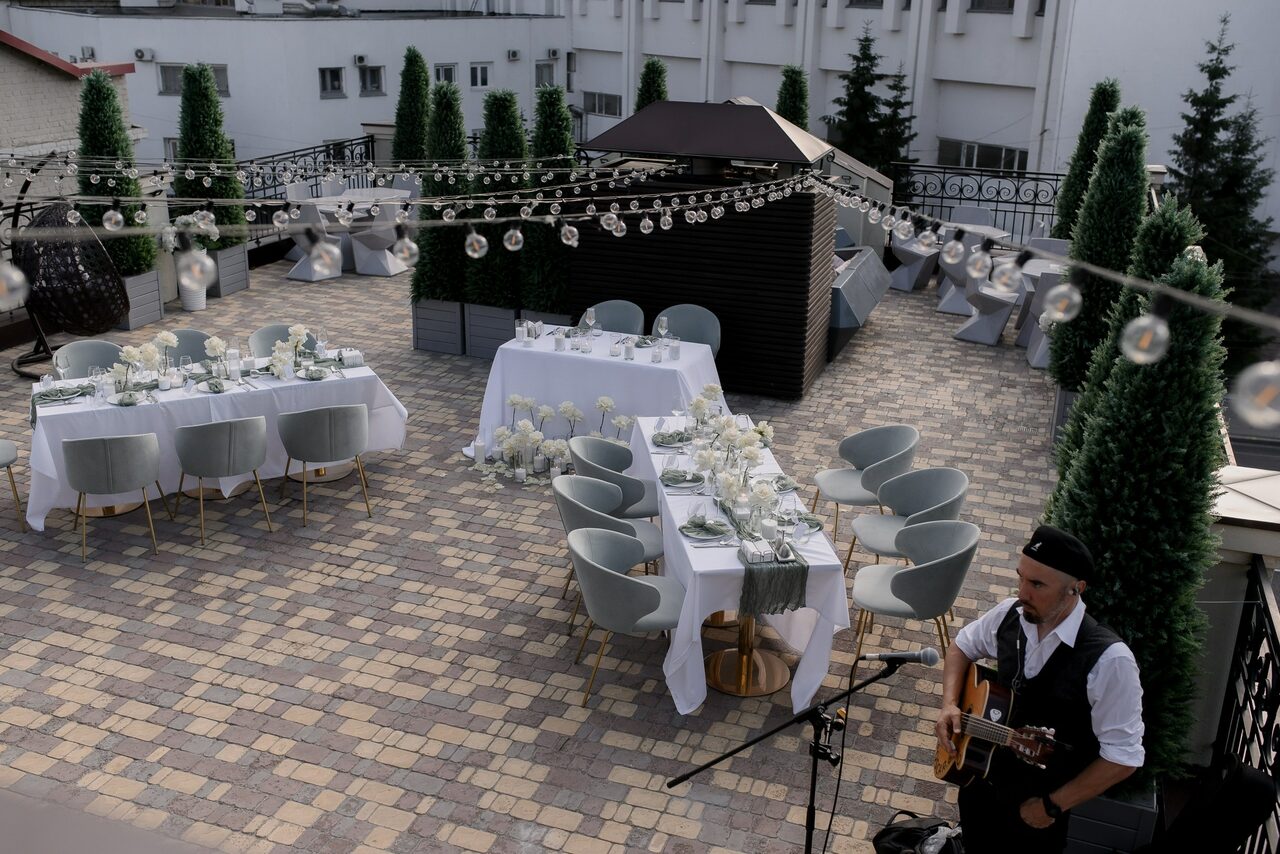 Арендовать Ресторан Небо Казани. Открытая терраса с брусчаткой, накрыты столы с белыми скатертями и серыми стульями. Декорирована гирляндами с лампочками. В углу играет гитарист. Расставлены высокие зеленые растения в горшках. На фоне белые стены здания.