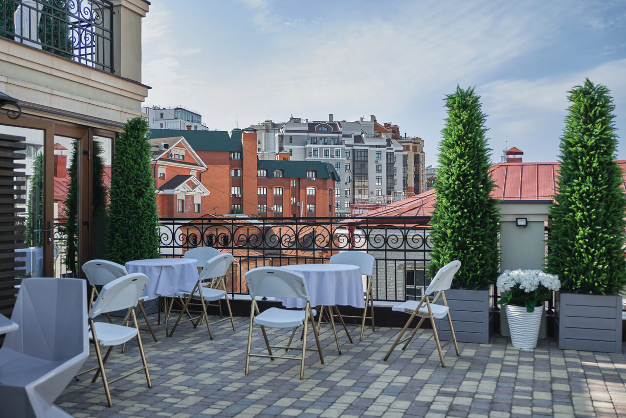 Арендовать Ресторан Небо Казани. Терраса с видом на городские здания, выложенная плиткой. Несколько круглых столов с белыми скатертями окружены складными стульями. Зелёные декоративные растения в горшках вдоль ограждения. Спокойный светлый день.
