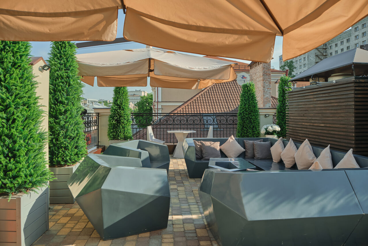 Арендовать Ресторан Небо Казани. Терраса с деревянным настилом, серыми геометрическими скамьями с подушками, столом, зонтиками, высокими зелеными растениями в горшках. Виден металлический забор, крыши и здания вокруг. Хорошая освещенность.