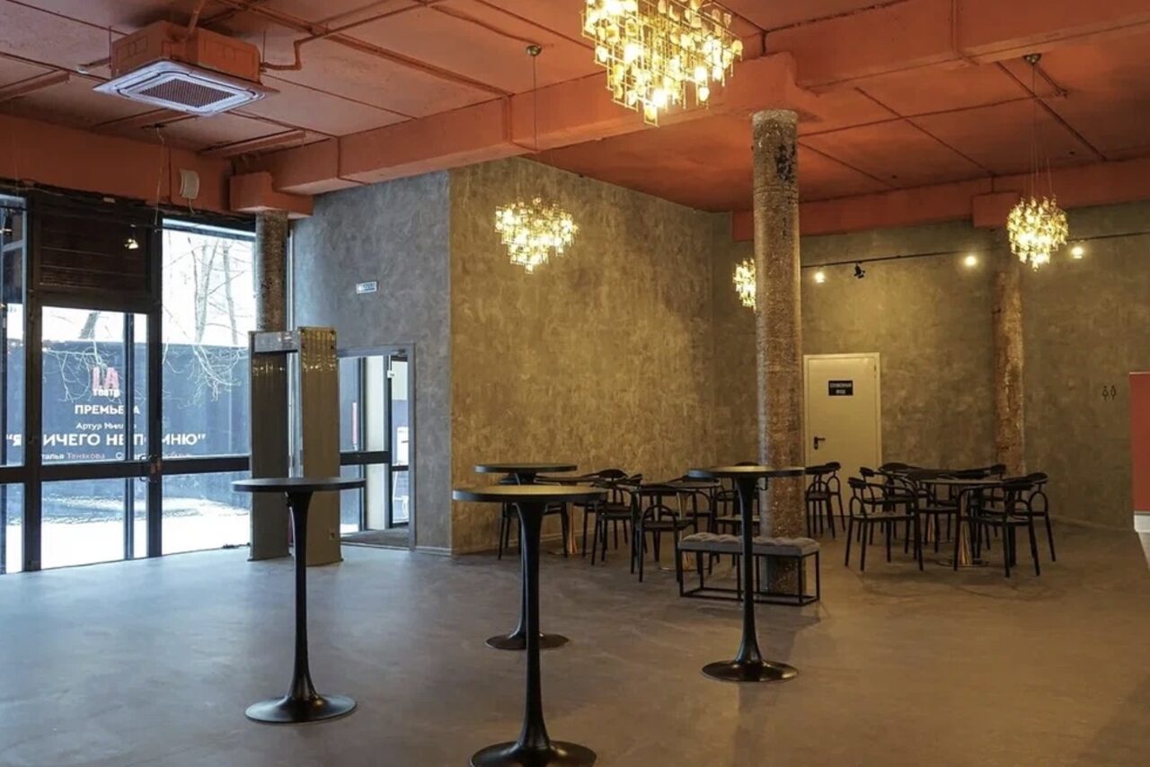 Арендовать Театр в Хамовниках. Просторное помещение с выполненными в индустриальном стиле стенами, равномерно расположенными столиками и стульями. Выделяются стеклянные двери, декоративные светильники и ярко оранжевый потолок с видимой вентиляцией.