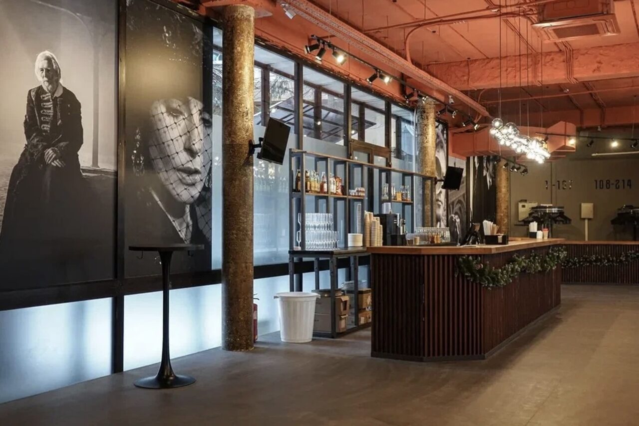 Арендовать Театр в Хамовниках. Барное пространство в индустриальном стиле с бетонными колоннами и деревянной стойкой. Большие чёрно-белые портреты украшают стены. На потолке открытые коммуникации. Освещение представлено подвесными лампами. Полки с бутылками, мусорная корзина.