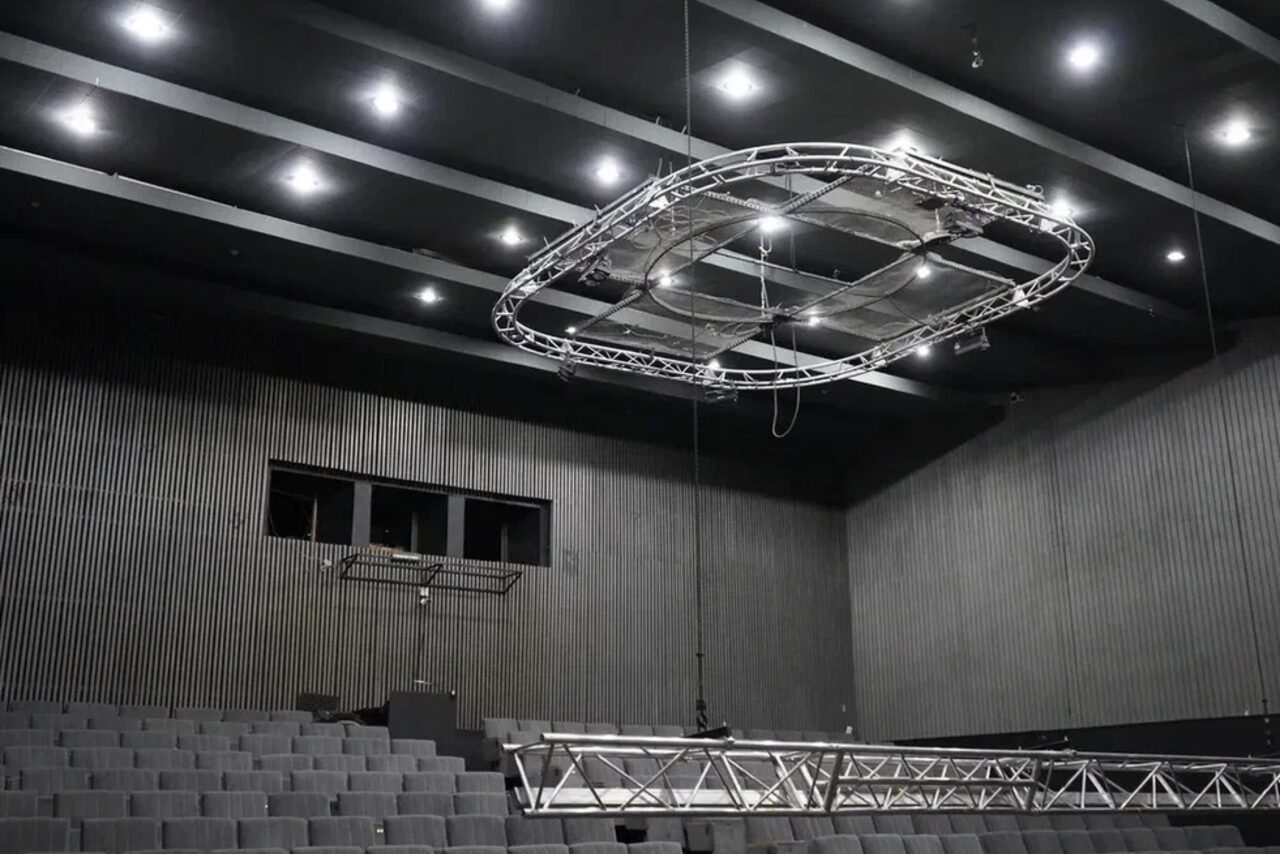 Арендовать Театр в Хамовниках. Зал с амфитеатром, серые ступенчатые ряды кресел, тёмный рельефный потолок с лампами, подвесная металлическая конструкция, стены отделаны вертикальными панелями серого цвета.