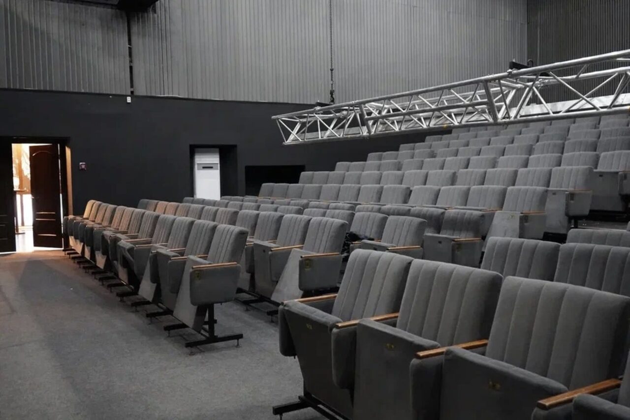 Арендовать Театр в Хамовниках. Аудиторный зал с рядной расстановкой серых кресел с подлокотниками. Пол покрыт ковролином, стены темные. Над креслами металлическая конструкция для освещения. Справа от двери виден шкаф. Освещение из дверного проема.