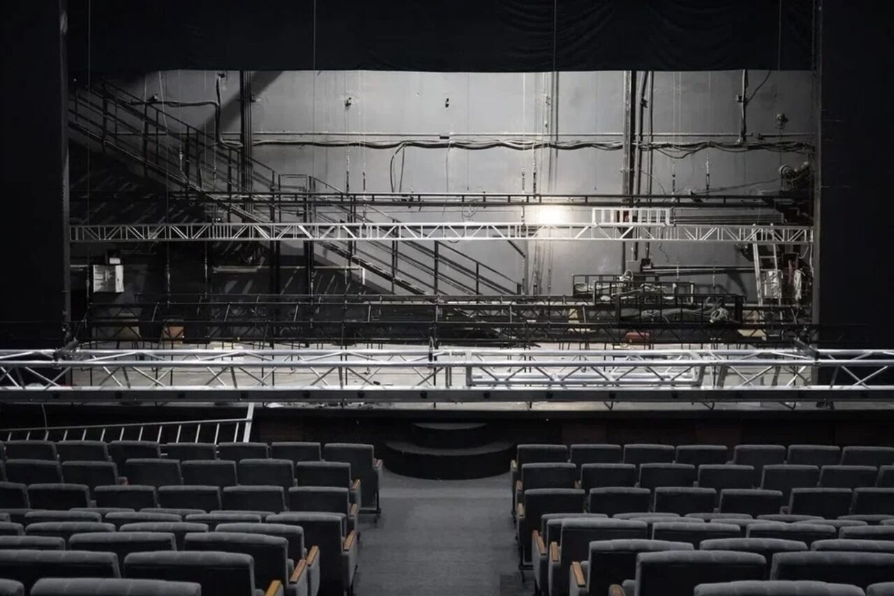 Арендовать Театр в Хамовниках. Аудитория со сценой и рядами темных кресел. На сцене установлена металлическая конструкция. Задняя стена с серыми панелями и трубами. Освещение тусклое, фон серый.