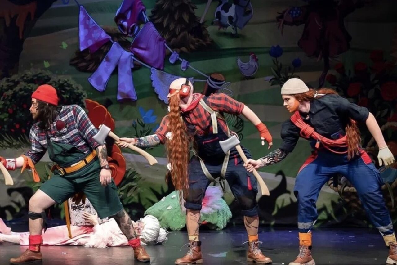 Арендовать Театр в Хамовниках. Театральная сцена с декорациями леса на заднем плане. Трое мужчин в театральных костюмах лесорубов с топорами. Яркие наряды, многоцветное освещение. На сцене лежат декоративные элементы, включая ткани и искусственную траву.