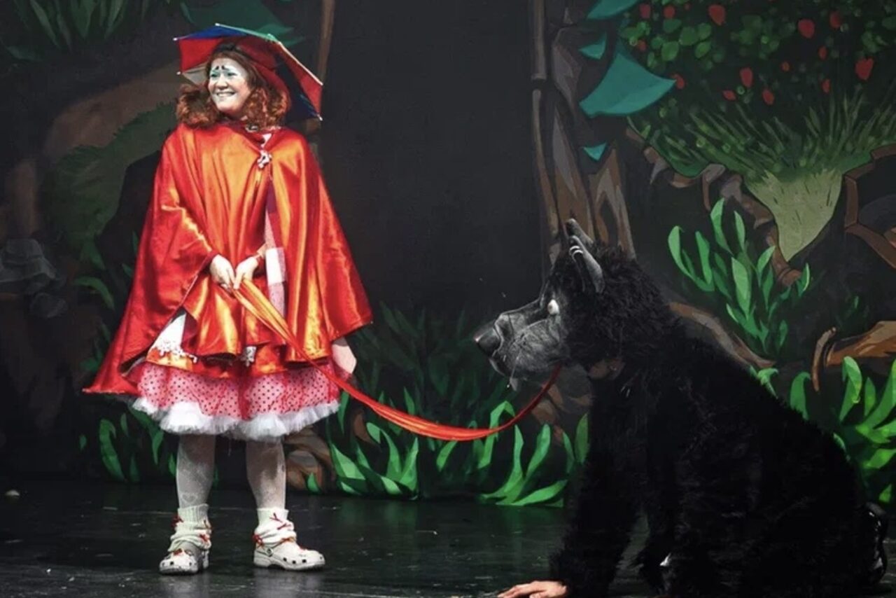 Арендовать Театр в Хамовниках. Выполняется театральное представление. Женщина в красном костюме с капюшоном на сцене с искусственно оформленным задником в виде леса. На сцене также персонаж в чёрном костюме волка, которого держат на поводке.