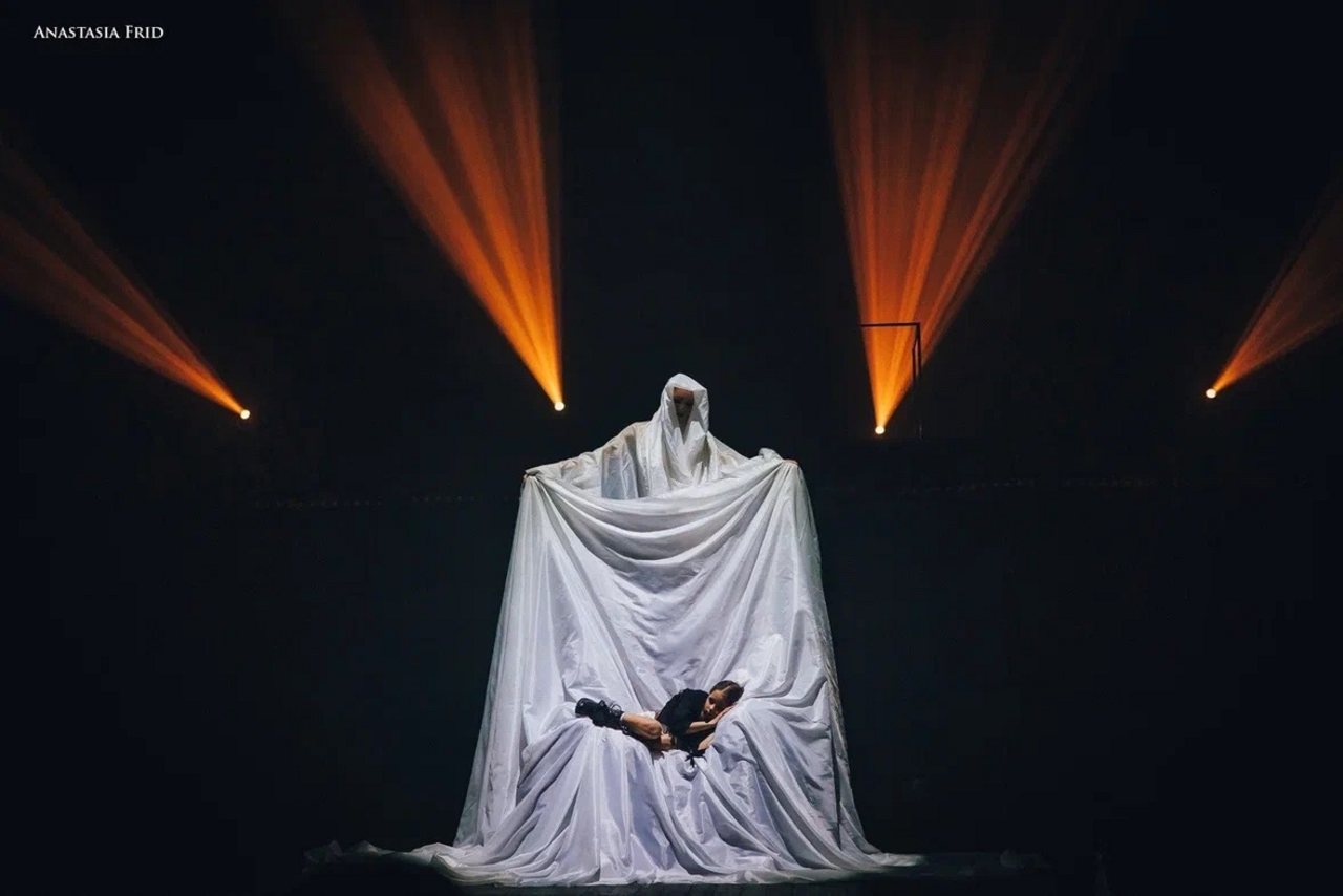 Арендовать Театр в Хамовниках. ALT: Темное сценическое пространство с чёрным фоном, на котором яркие прожекторы создают световой эффект. В центре крупная драпировка из белой ткани, на которой выступают два человека, один из которых поднимает ткань.