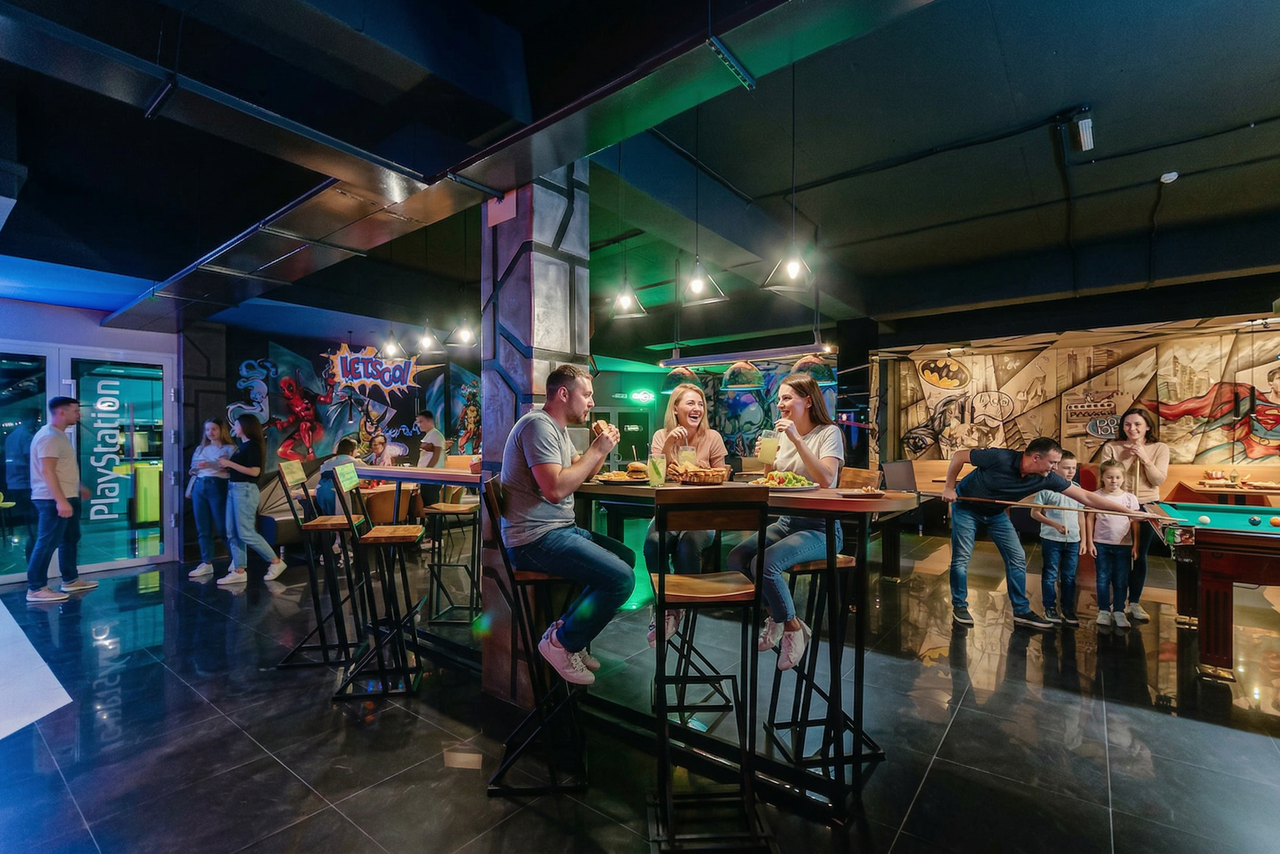 Арендовать Площадка для праздников Spark. Пространство включает зону с высокими столами и стульями, бильярдный стол. Яркое освещение, темные полы. На стенах яркие граффити, изображены персонажи. Надпись "PlayStation". Композитный материал мебели, яркие цвета в интерьере.