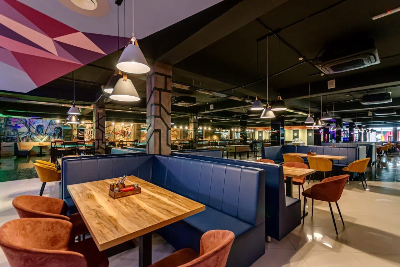 Арендовать Площадка для праздников Spark. Просторное помещение с деревянными столами и синими диванами-кабинами. Коричневые стулья с мягкой обивкой. Современные подвесные светильники. Пол из светлого материала. Стены с граффити. Открытые коммуникации на потолке.