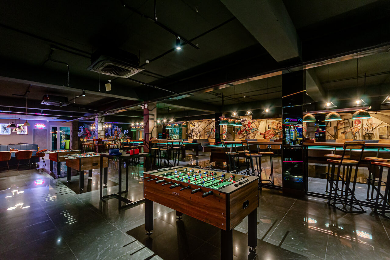 Арендовать Площадка для праздников Spark. Современное помещение для досуга с настольным футболом, барной стойкой и стульями. Интерьер выполнен в тёмных тонах, на стенах - абстрактный арт. Пол блестящий, освещение точечное, подвесное. Видна вывеска неоновая и телевизор на стене.