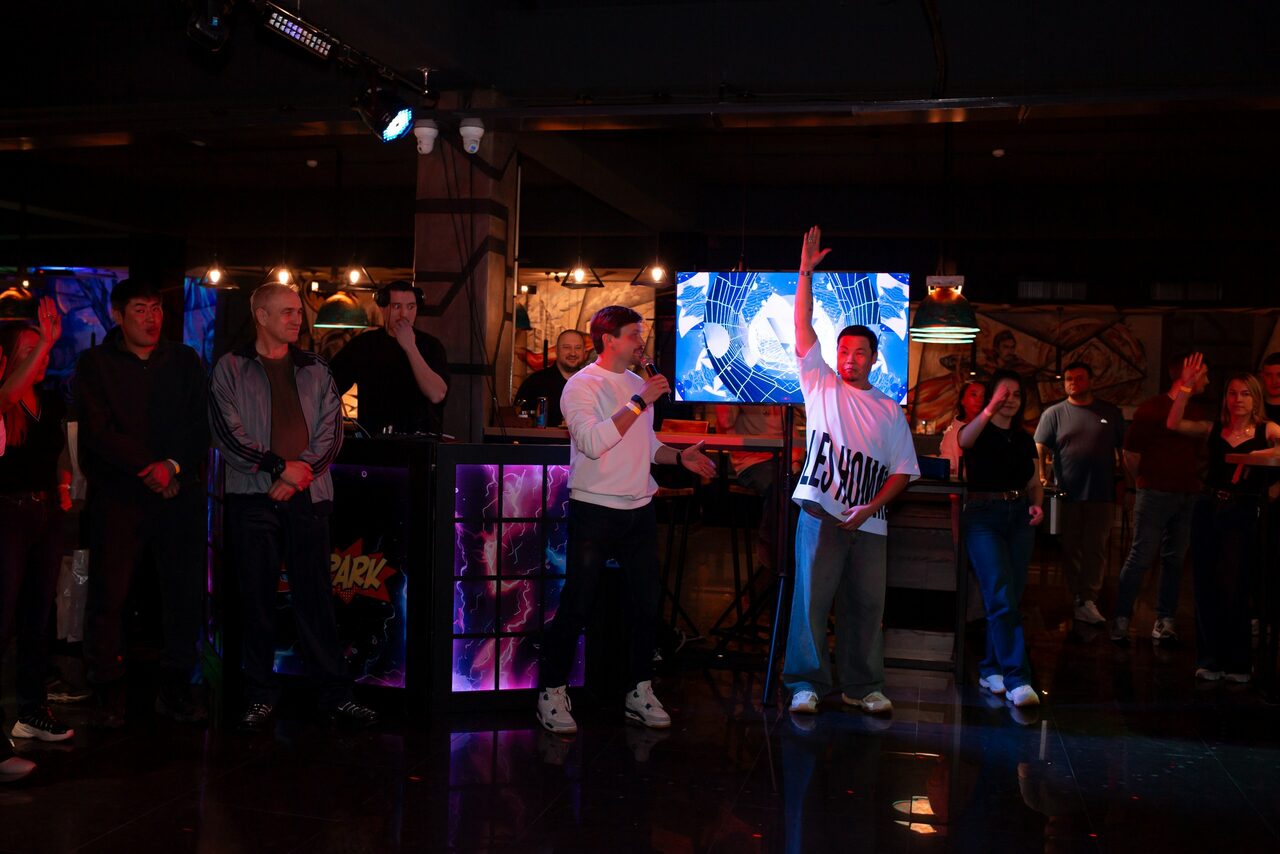 Арендовать Площадка для праздников Spark. Изображение интерьера с темным освещением, искусственные лампы создают разноцветное освещение. На переднем плане группа людей, один человек держит микрофон. На стене экран с яркими изображениями. Пол отражает свет.