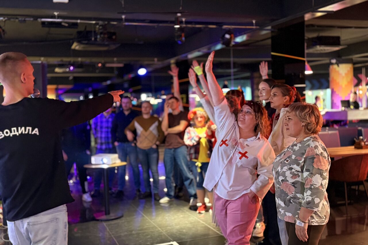 Арендовать Площадка для праздников Spark. Современное помещение с темным потолком и полом, напряженной атмосферой. Группа людей стоит вокруг, некоторые подняли руки. На переднем плане доска или стол. Яркое разноцветное освещение. На человеке чёрная футболка с надписью.