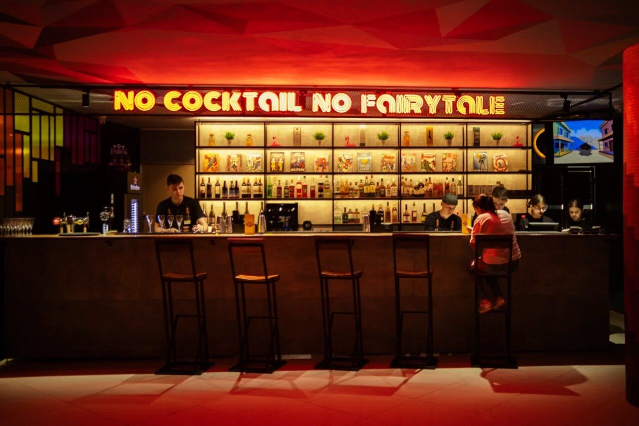 Арендовать Площадка для праздников Spark. Барное пространство с длинной стойкой, пять высоких стульев, стеллажи с бутылками, неоновая надпись "No Cocktail No Fairytale". Освещение теплое, цветовая гамма включает красный и черный, на стене декоративные элементы.