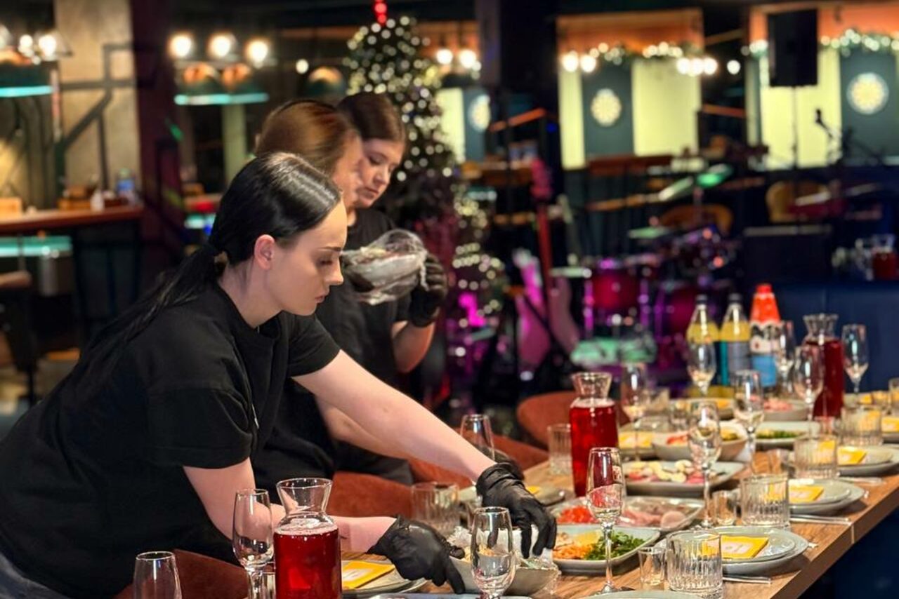 Арендовать Площадка для праздников Spark. Изображение большого зала ресторана, оформленного в праздничном стиле. Накрытые столы с блюдами, напитками и приборами. Три человека сервируют столы. На заднем плане видны элементы освещения и барабанная установка.