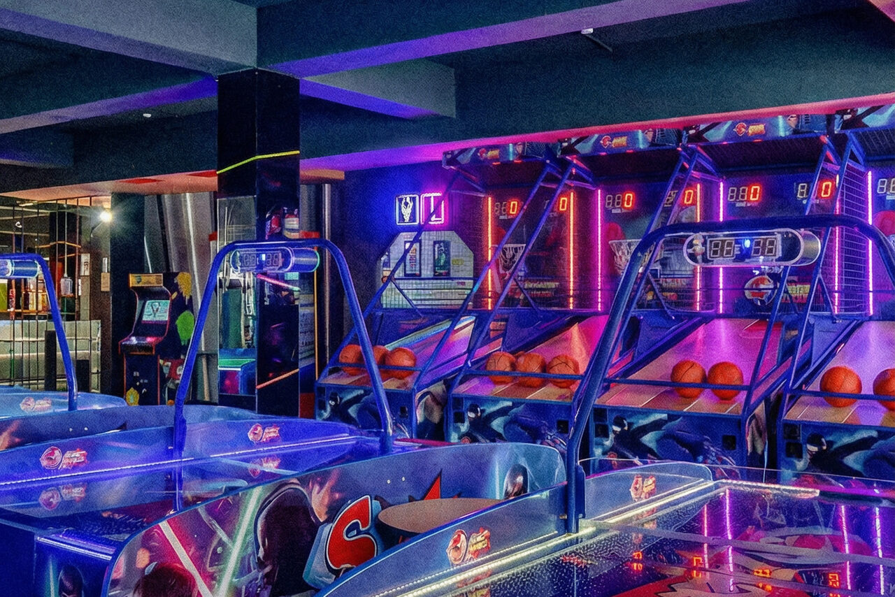 Арендовать Площадка для праздников Spark. Игровое пространство с несколькими баскетбольными автоматами, яркой неоновой подсветкой синего и фиолетового цветов. На стене расположены экраны со счетами. Различные игровые автоматы видны на заднем плане.