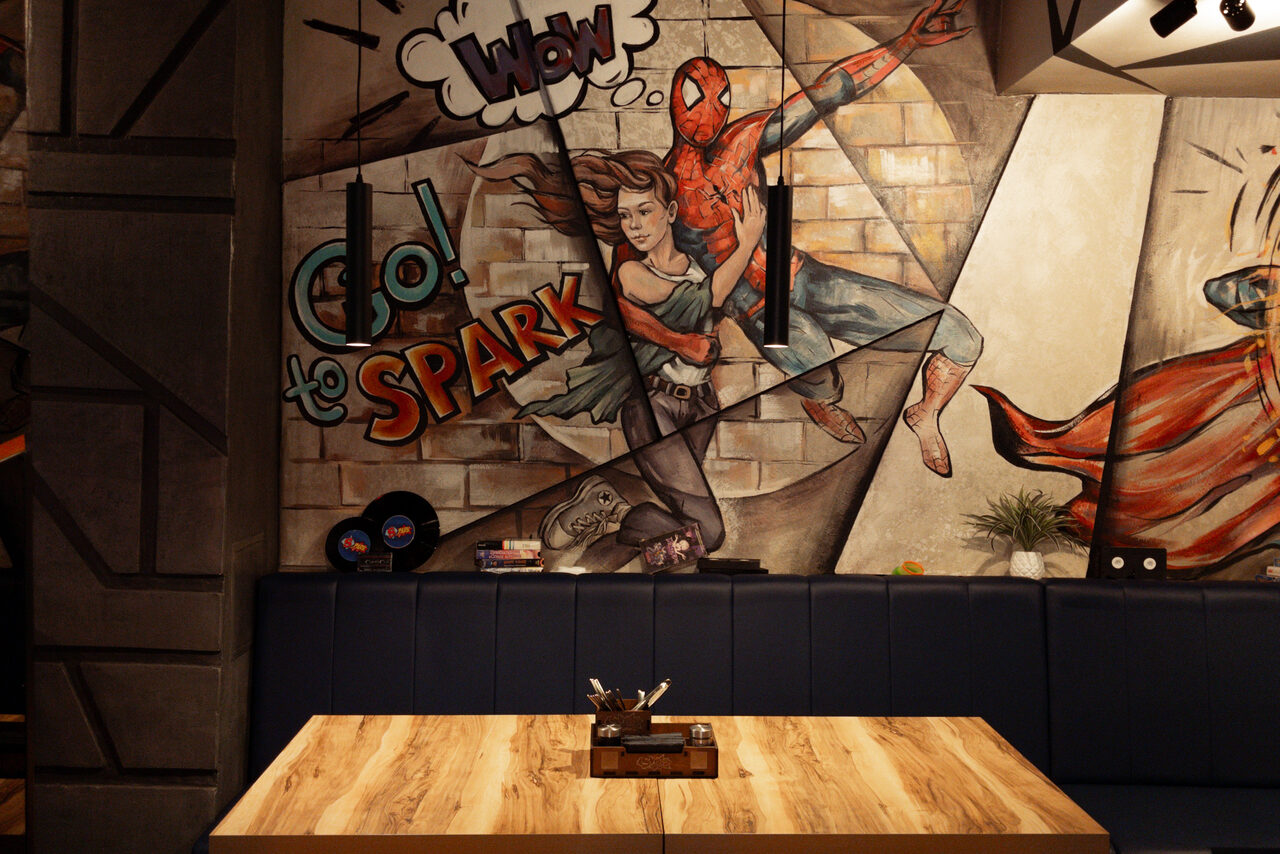 Арендовать Площадка для праздников Spark. Настенное граффити с супергероем и надписями на кирпичной стене. Деревянный стол с посудой, окружён синим мягким диваном. Потолок с геометрическими светильниками. Комната освещена теплым светом. На заднем плане декоративные элементы.