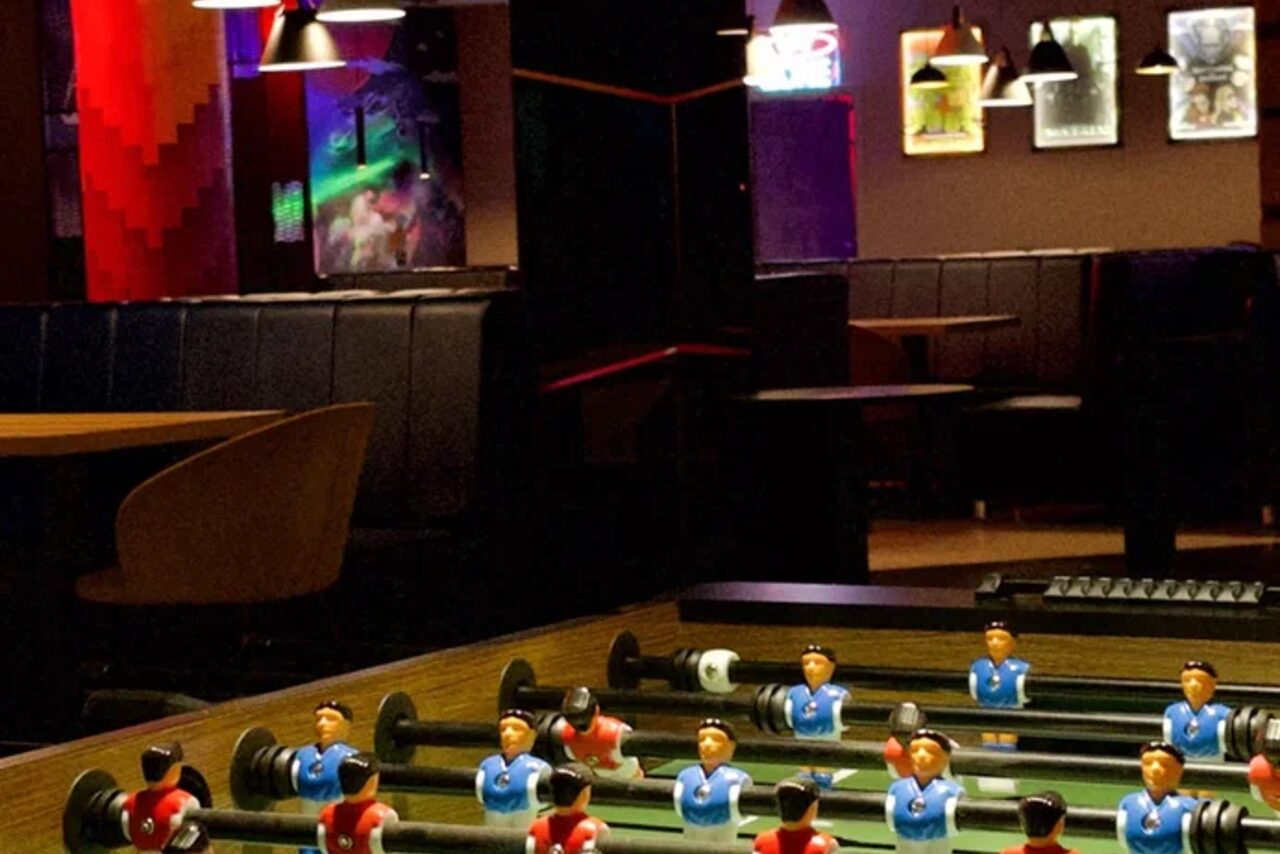 Арендовать Площадка для праздников Spark. Открытое помещение с настольным футболом. На заднем плане кожаные диваны, деревянные столы и кресла. Стены украшают цветные постеры. Освещение от подвесных ламп. Светлые и темные оттенки коричневого с яркими акцентами.