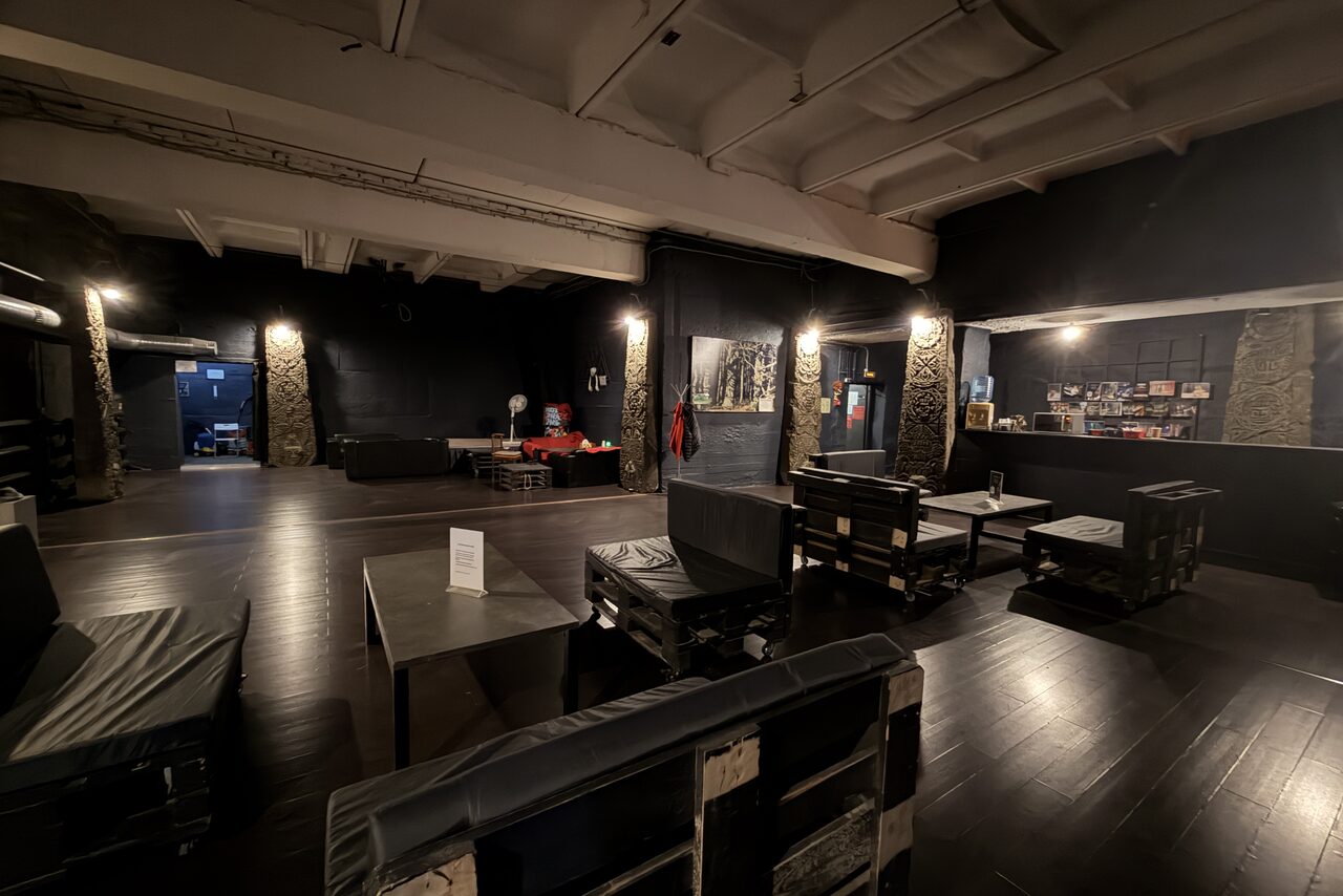 Арендовать Монолит Холл. Open loft-style space with dark wood floors and black walls. Pallet seating with cushions, central tables, and bar area. Industrial lighting with ceiling fixtures and wall sconces. Exposed pipes and textured columns. Art and decor on walls.