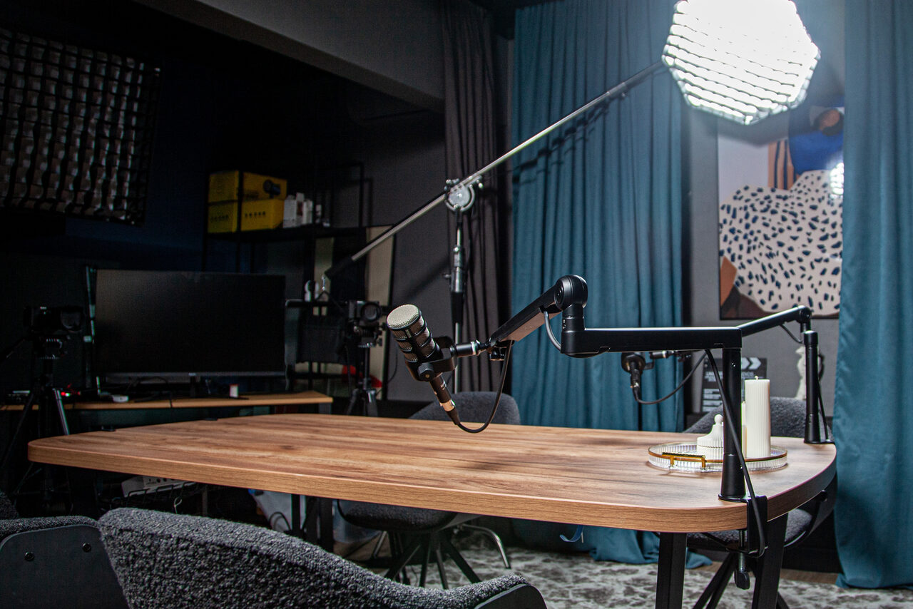Арендовать Студия для записи подкастов "Искусство Подкаста". Подкаст-студия с деревянным столом, на котором установлены микрофоны на стойках. Стулья вокруг стола, световое оборудование. Фон оформлен шторами. Присутствует монитор, черные полки с техникой и коробками. Геометрический постер на стене.
