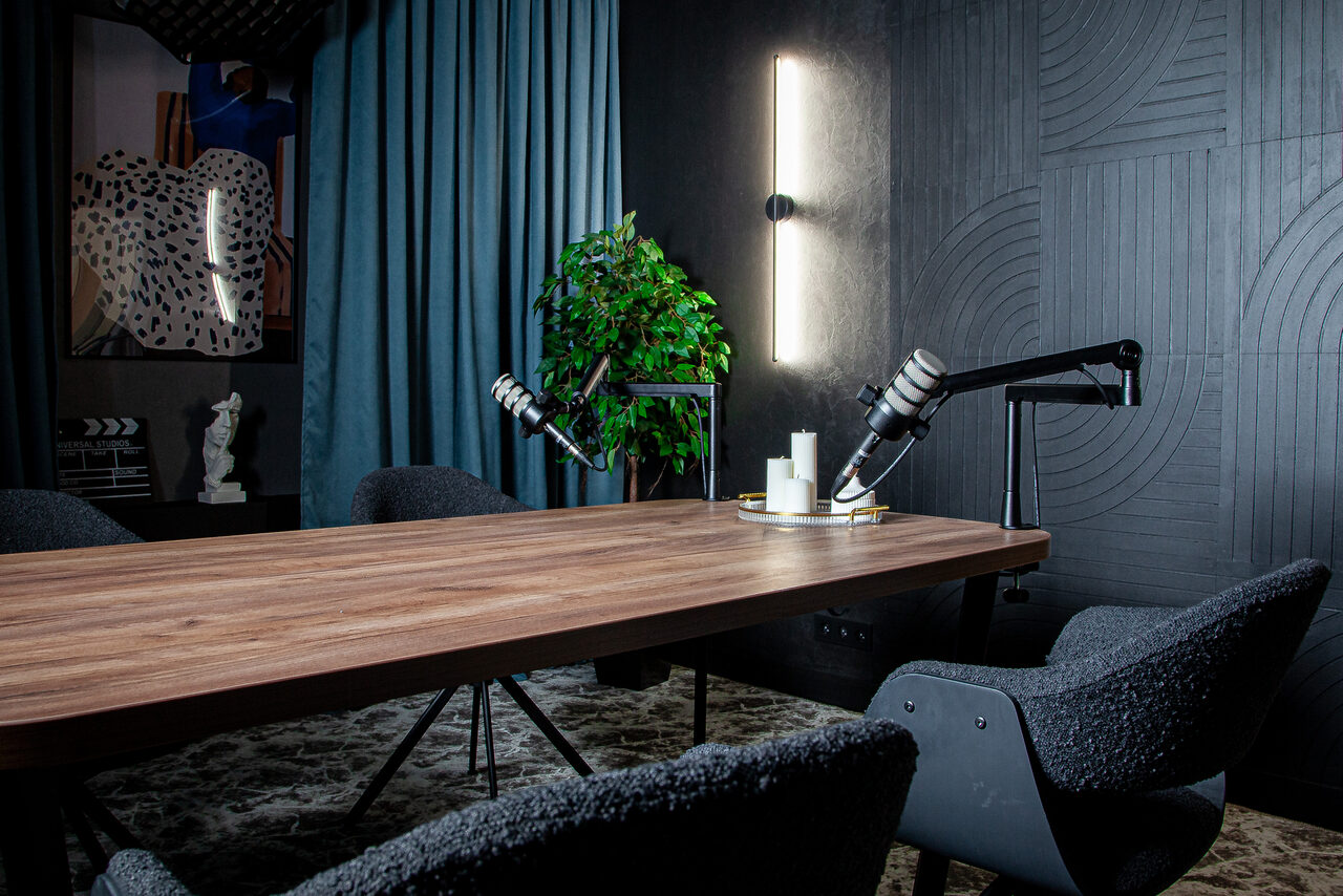 Арендовать Студия для записи подкастов "Искусство Подкаста". Комната с длинным деревянным столом и тремя темными креслами. На столе два микрофона на стойках. Справа вертикальный светильник на стене. На заднем плане синие шторы, зелёное растение и стены с черным рельефным узором.