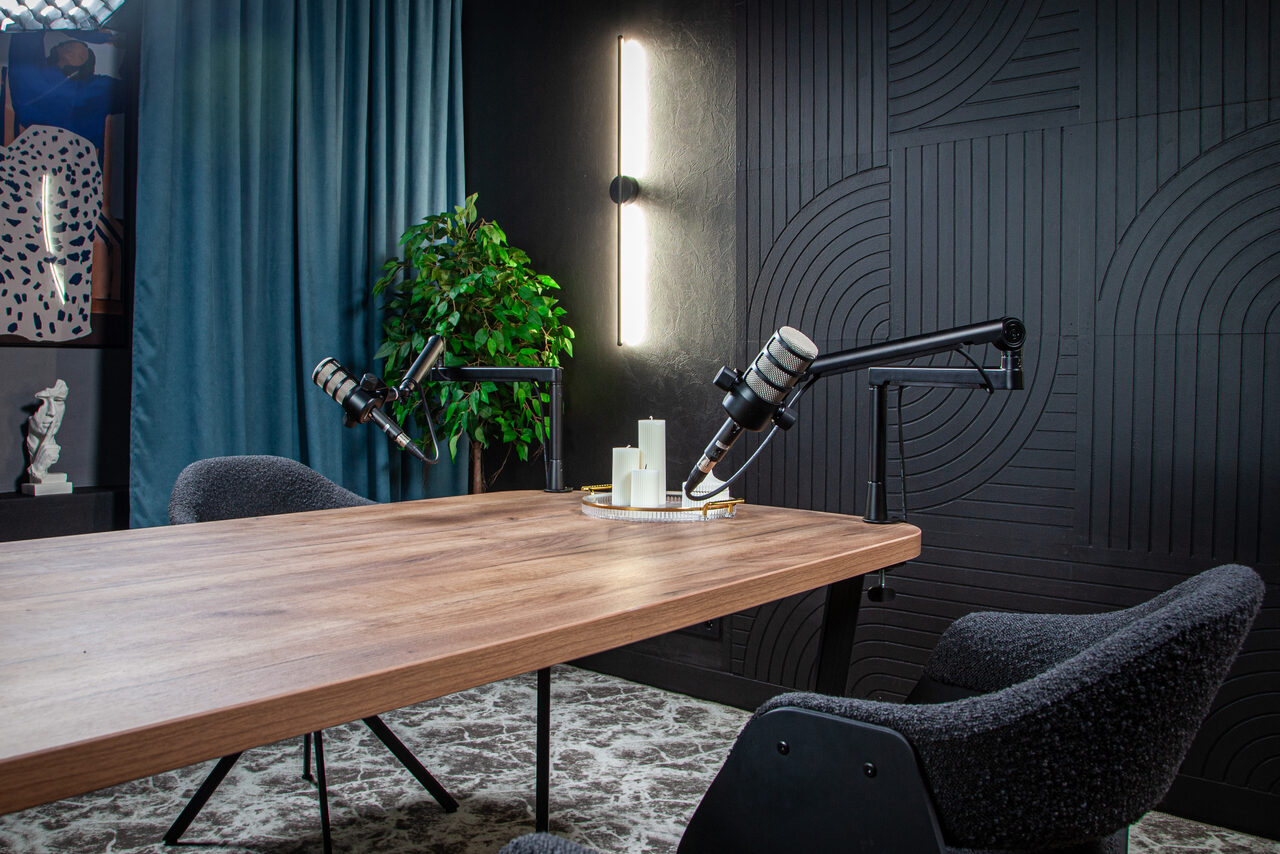 Арендовать Студия для записи подкастов "Искусство Подкаста". Современная студия с деревянным столом, оборудованным двумя микрофонами на стойках. Уютные серые кресла, ковёр, декоративные панели на стене, вертикальное освещение на чёрной стене, синий занавес и зелёное растение.