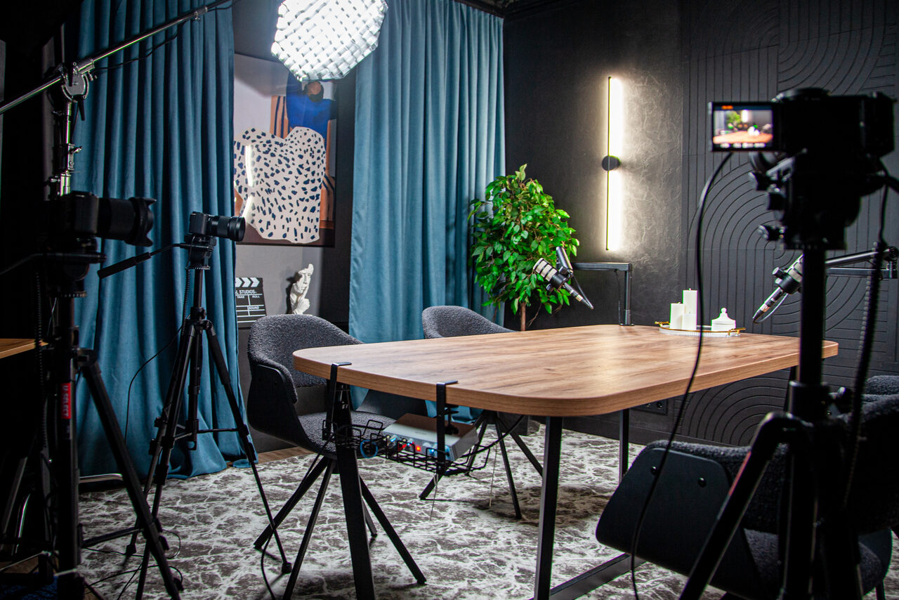 Арендовать Студия для записи подкастов "Искусство Подкаста". Современное студийное пространство с деревянным столом и серыми креслами. Съемочное оборудование, включая камеры и свет. Темные стены, голубые шторы, зеленое растение. Вертикальный светильник на стене. Настенный постер и акустическая панель.