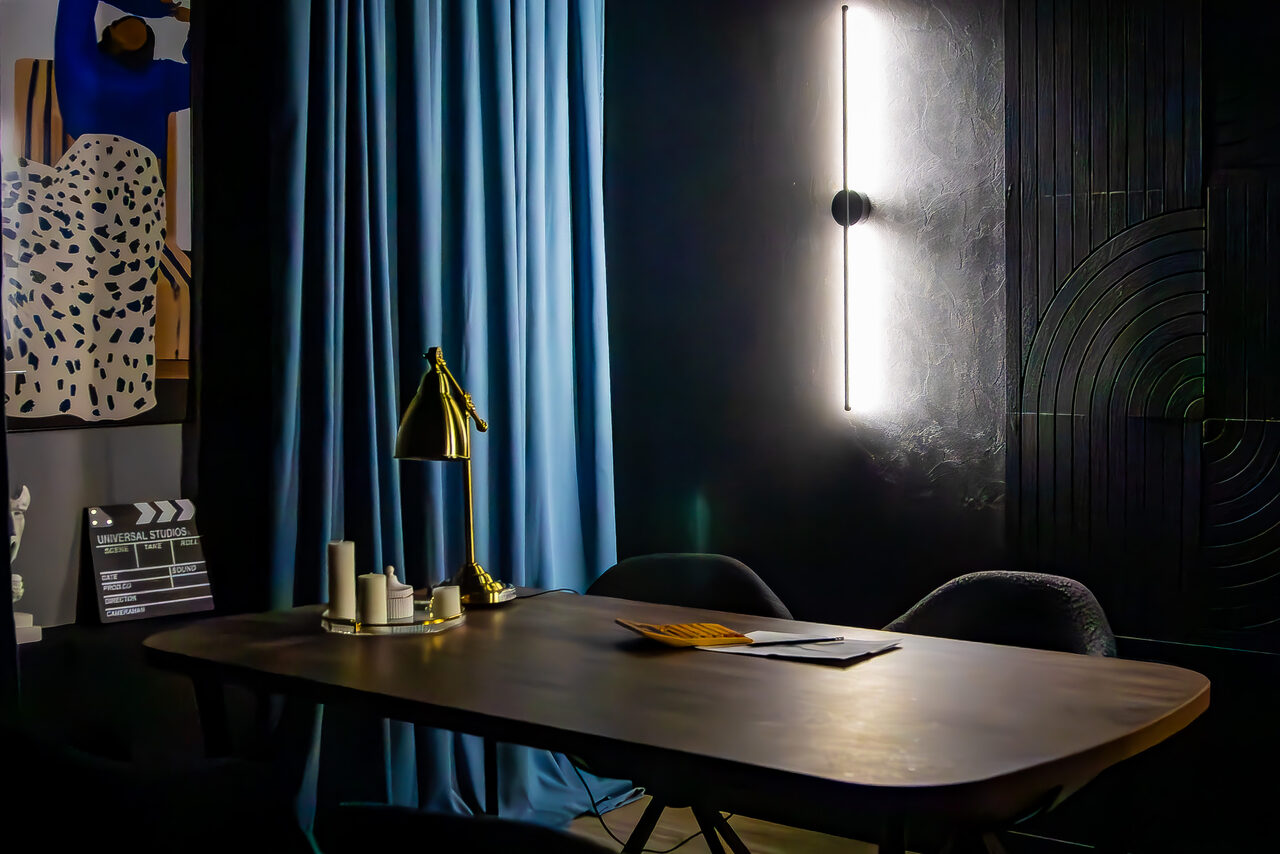 Арендовать Студия для записи подкастов "Искусство Подкаста". Комната с тёмными стенами, деревянным столом и парой чёрных кресел. Вертикальная настенная лампа, голубая штора, настольная лампа золотистого цвета. На столе свечи и документы. На стене фото с абстракцией и чёрно-белая хлопушка.