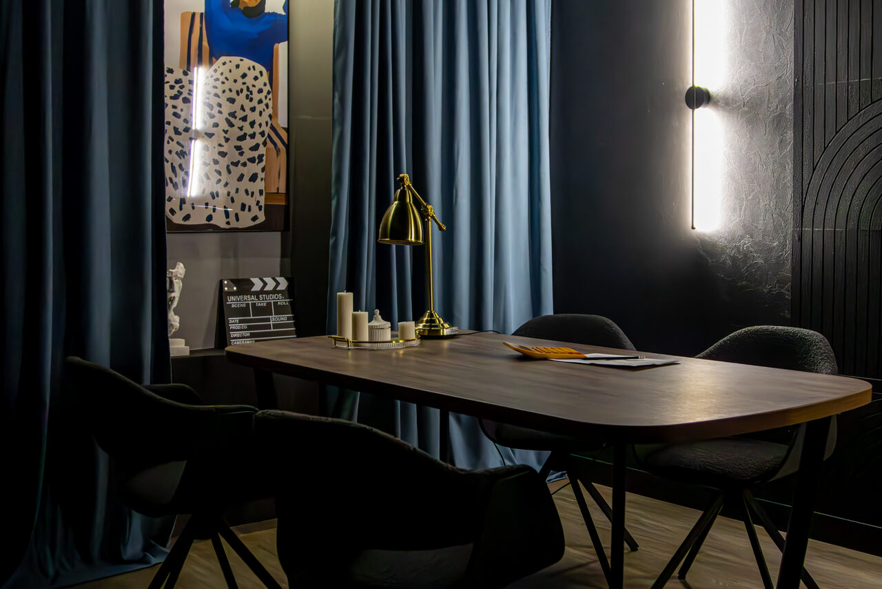 Арендовать Студия для записи подкастов "Искусство Подкаста". Современное офисное пространство с темным деревянным столом и четырьмя мягкими стульями. На столе лампа, свечи и книги. Синие шторы, настенные светильники, темные стены с декоративными элементами. На стене постер и классическая мраморная скульптурка.