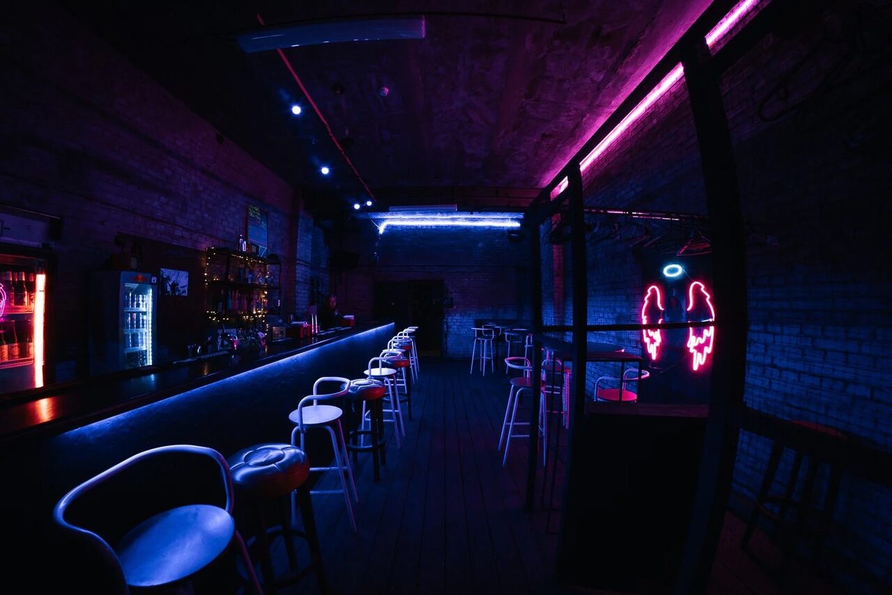 Арендовать PVZ CLUB (ПВЗ Клуб). Барная зона с длинной стойкой и высокими стульями. Кирпичные стены, неоновые огни с оттенками синего и розового. На стене неоновая фигура крыльев с ореолом. Холодильник и полки с напитками. Пол тёмный, потолок индустриального стиля.