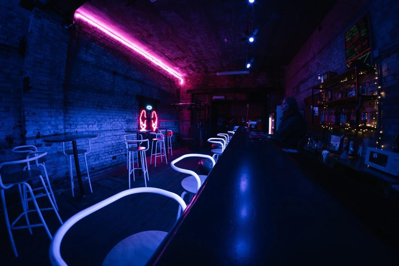 Арендовать PVZ CLUB (ПВЗ Клуб). Клубное пространство с длинной стойкой и высокими стульями, кирпичные стены, неоновая подсветка фиолетового и синего цвета, полки с напитками, таблички с меню на стене, приглушенное освещение.