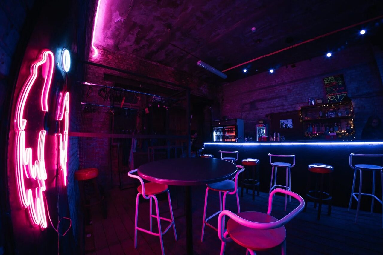 Арендовать PVZ CLUB (ПВЗ Клуб). Интерьер барного пространства с неоновым освещением. Круглый стол и стулья в центре, барная стойка с высокими стульями справа. На стене неоновая вывеска. Цвета: розовый, синий. Потолок и стены из кирпича, пол из дерева. Светодиодные лампы на стенах.