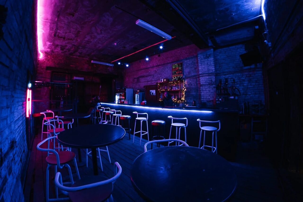 Арендовать PVZ CLUB (ПВЗ Клуб). Барное помещение с кирпичными стенами, темным деревянным полом и неоновым голубым и розовым освещением. Барная стойка с высокими стульями, круглые черные столы с белыми стульями, светильники на потолке.