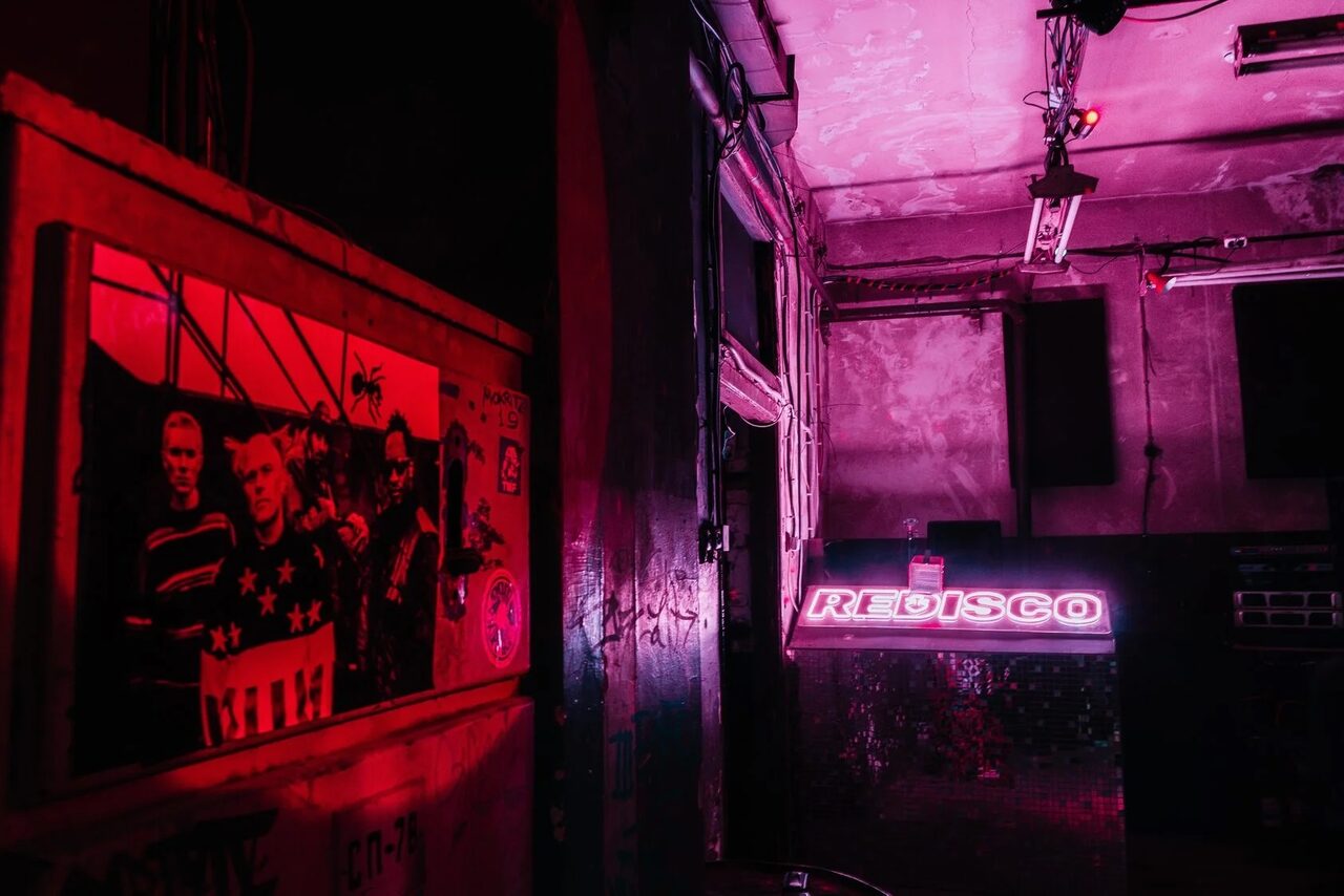 Арендовать PVZ CLUB (ПВЗ Клуб). Тёмное пространство с неоном, барная стойка с вывеской "DISCO", стены с граффити, потолок с обшарпанной краской, тусклое освещение красного оттенка, на стенах плакаты, в том числе с изображениями людей, провода на потолке.