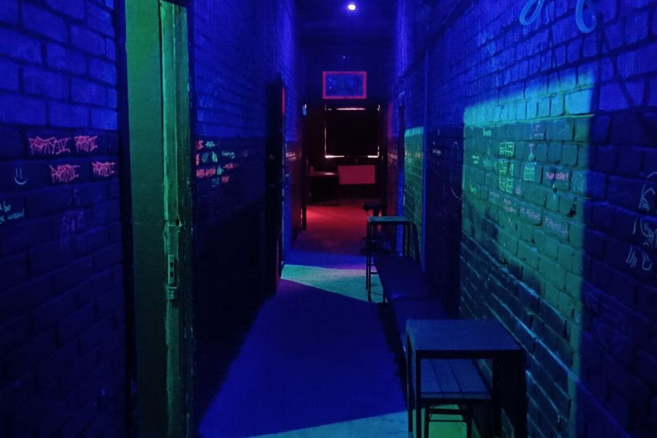 Арендовать PVZ CLUB (ПВЗ Клуб). Коридор с кирпичными стенами, освещенный синим и фиолетовым светом. На стенах разноцветные надписи. По правую сторону стоят металлические стулья и столы. Вдалеке видна более светлая комната с мебелью.