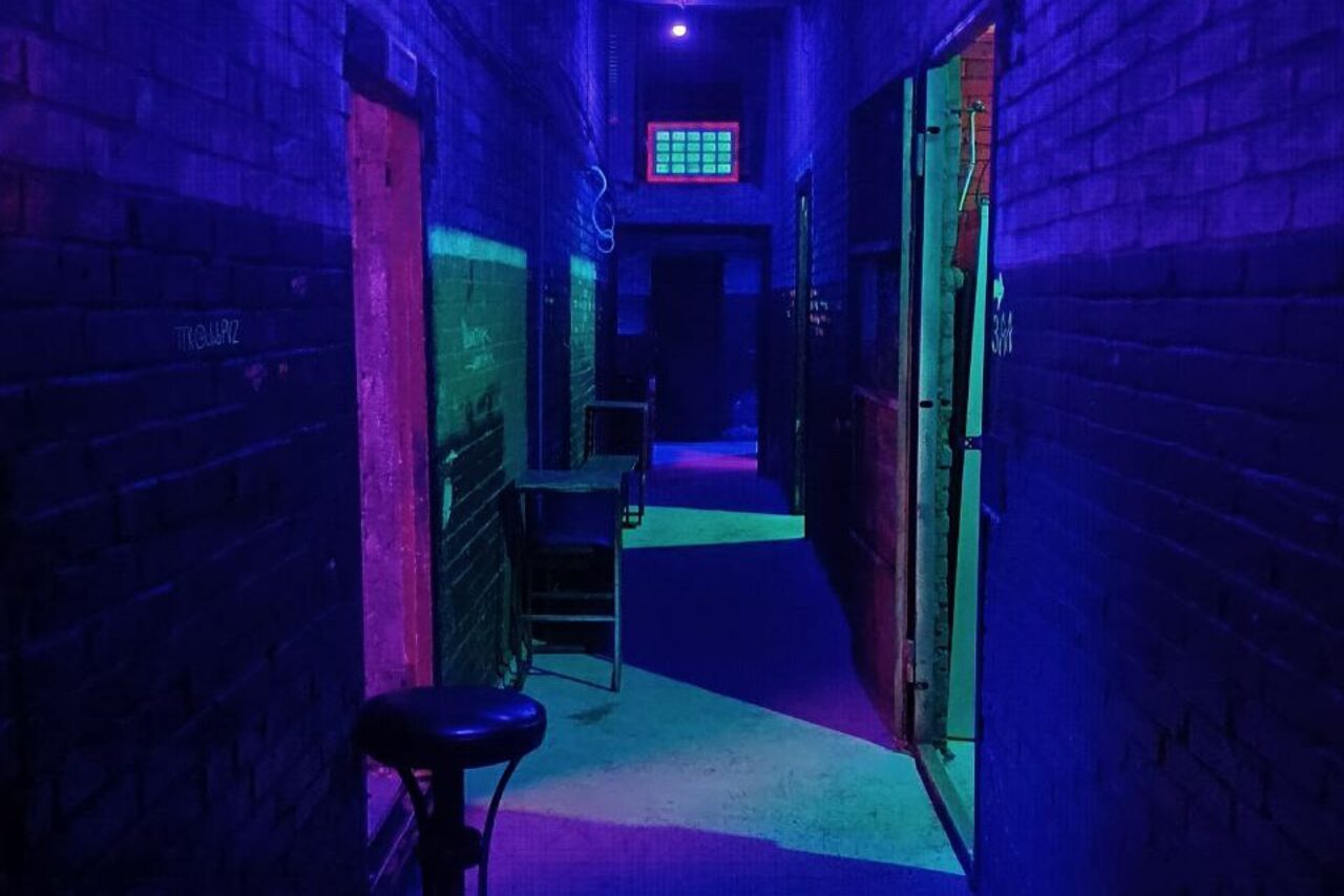 Арендовать PVZ CLUB (ПВЗ Клуб). Коридор с кирпичными стенами, освещенный фиолетовым и синим светом. Двери по обеим сторонам, слева стул и стол. Пол бетонный, в глубине небольшое квадратное окно с решеткой. На стене надписи.