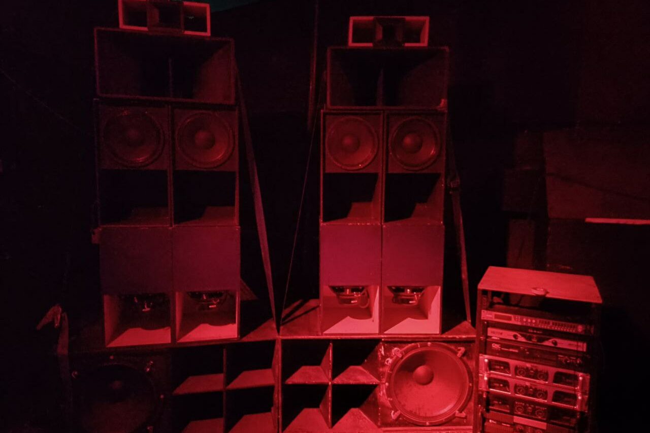 Арендовать PVZ CLUB (ПВЗ Клуб). Промышленное пространство с двумя большими аудиоколонками и оборудованием для звука. Освещение приглушённое, красное. Колонки и оборудование стоят рядом друг с другом.комплект в черно-красной цветовой гамме.