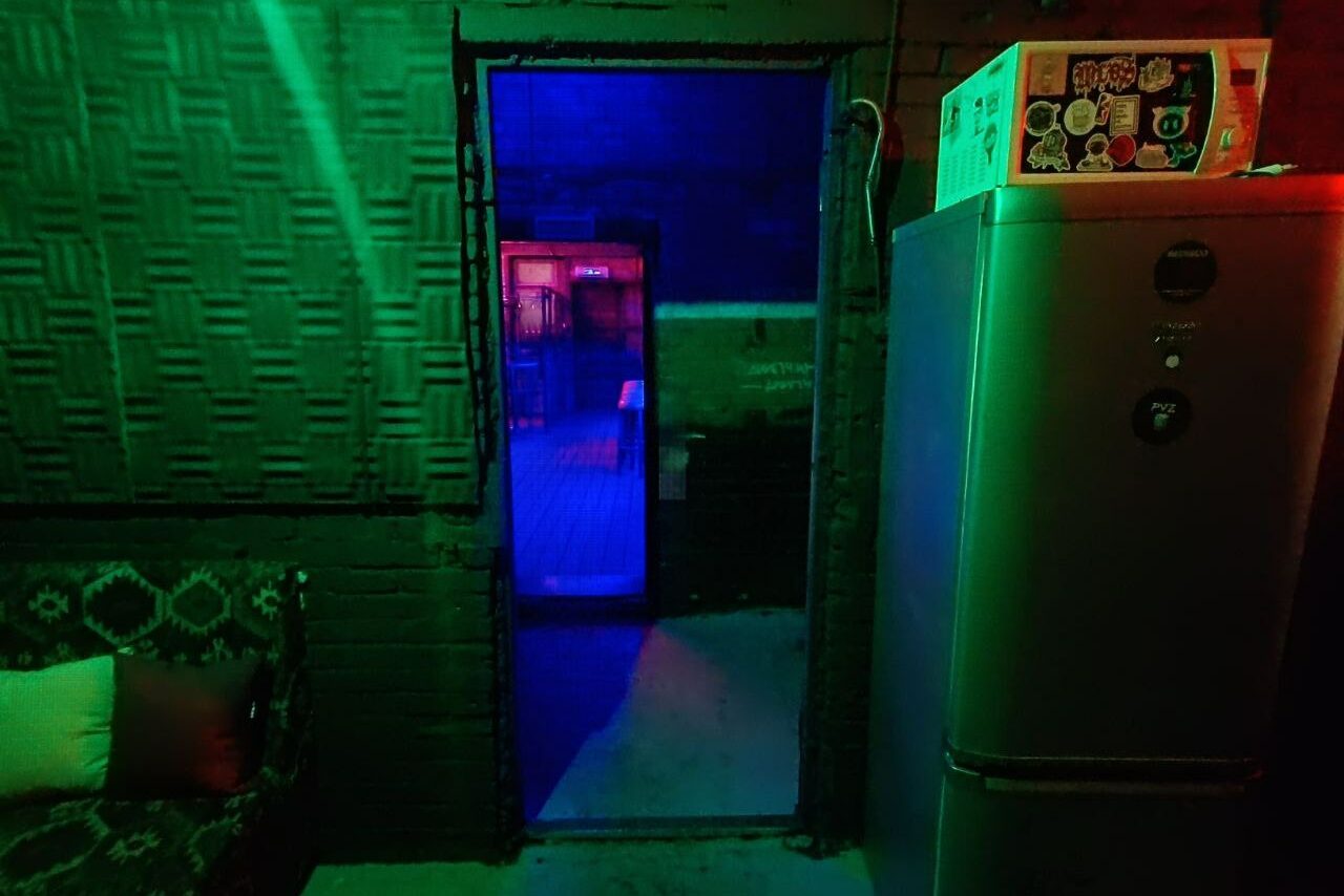 Арендовать PVZ CLUB (ПВЗ Клуб). Подсобное помещение с кирпичными стенами, обитое матами, полуоткрытая дверь ведет в соседнее помещение с красным и синим освещением. В комнате серый холодильник и диван с узором. Слабое зелёное освещение. На холодильнике коробка с наклейками.