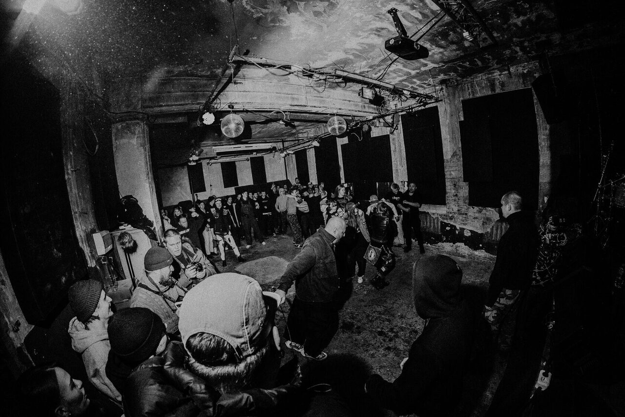 Арендовать PVZ CLUB (ПВЗ Клуб). Индустриальное пространство с бетонными стенами и потолком. Группа людей стоит вокруг свободной зоны. Потолочные балки с прожекторами и диско-шарами. Окна закрыты тёмными шторами. Незамысловатое освещение. Чёрно-белое изображение.